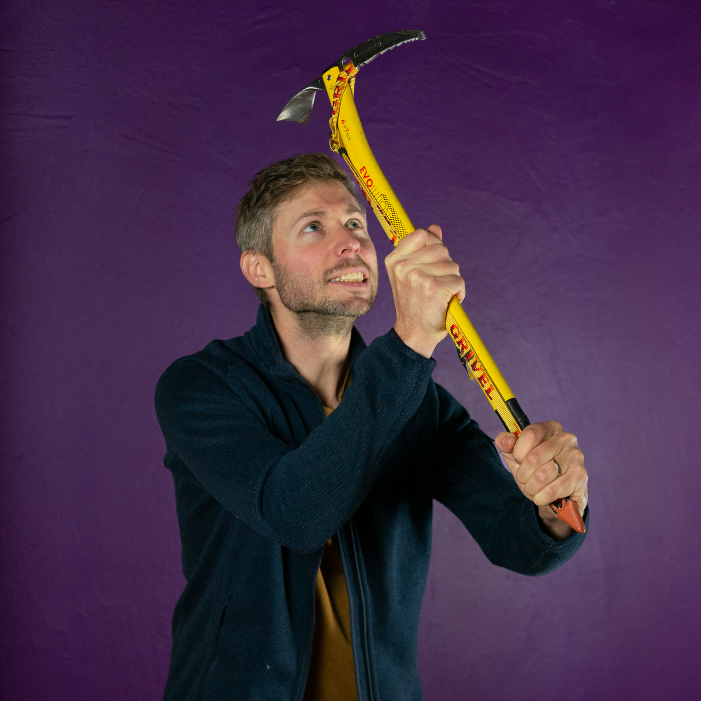A photo of Norman against a purple background. He's a white man with short dark hair and a beard and moustache. He's looking up to the sky and has an ice axe in his hands which he is wielding with both hands.