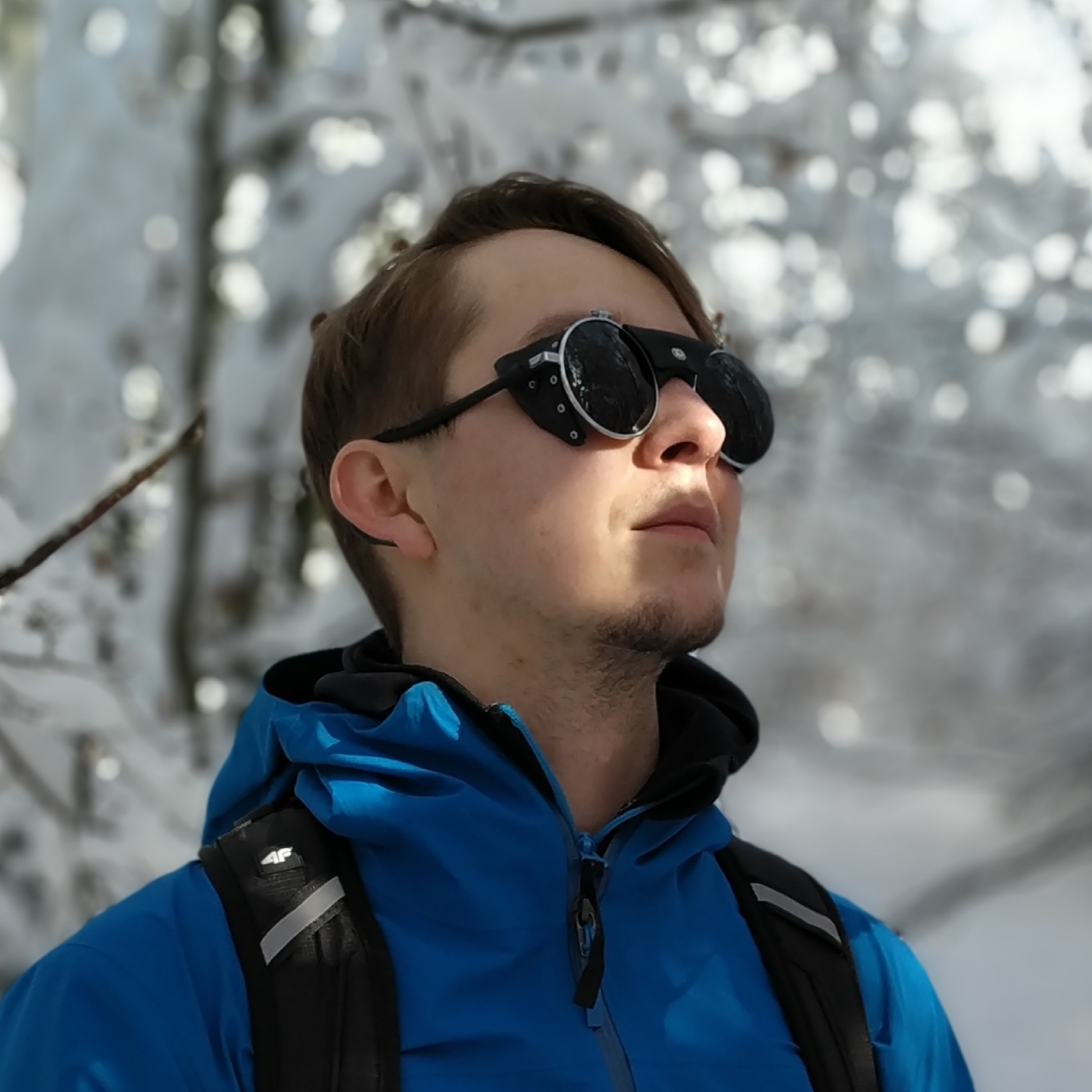 Photo of Patryk, a white man with dark brown short hair, wearing a dark blue coat with a black rucksack and with black circular ski goggles on. The background is showing snow covered trees and he's looking upwards to the right.