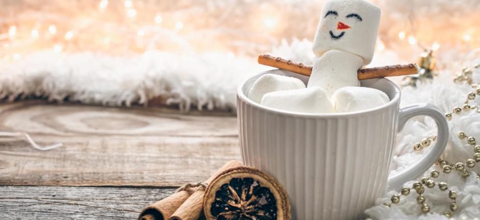 Photo of a mug containing hot chocolate with marshmallows, and some of the marshmallows have been made into the shape of a snowman with a face drawn on and hands and ears added. There are oranges and cinnamon on the table beside and behind is a blurred image of festive lights.