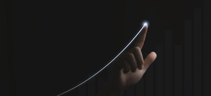 Photo of a dark room with a hand which is lit from below, index finger pointing toward the screen with a bright white light at the tip, which has traced a growth curve in light below and to the left corner.