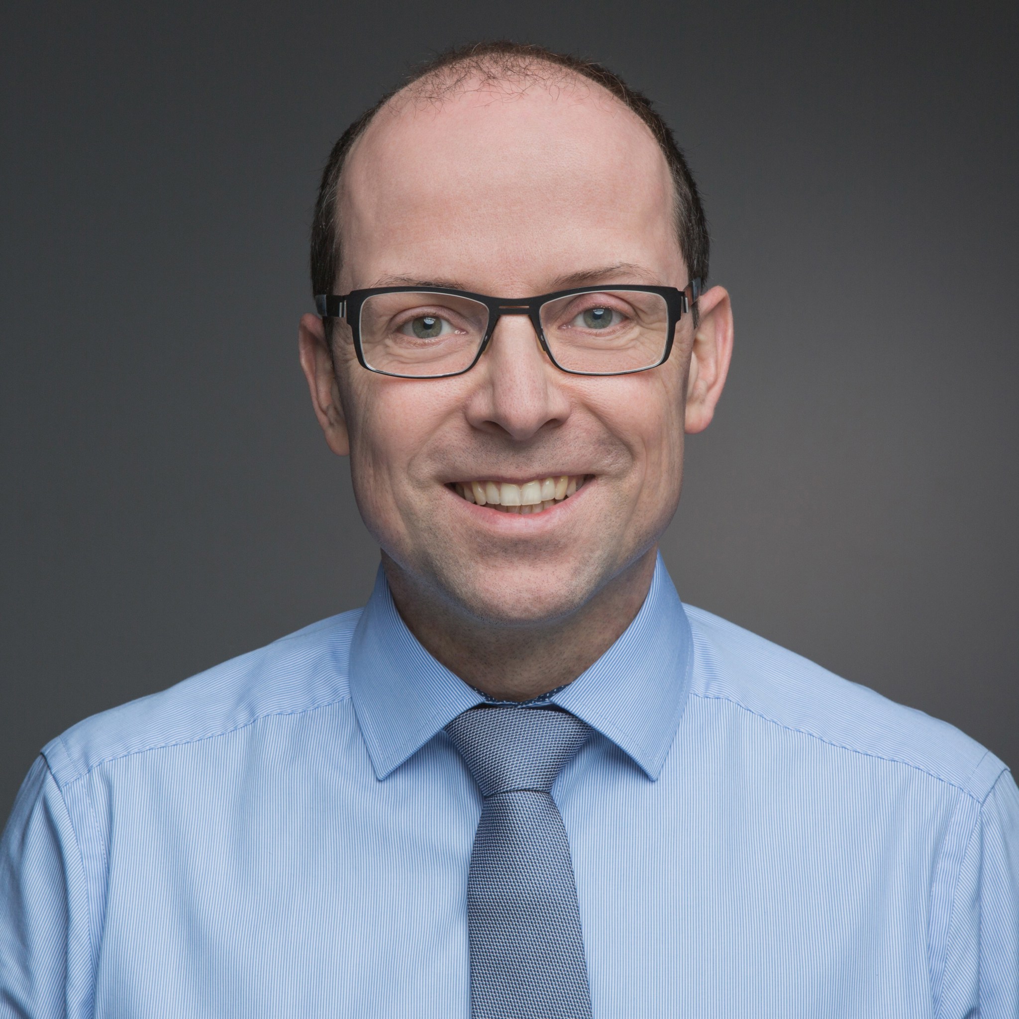 Photo of a white man looking at the camera. He's wearing glasses and has a bald head. He's wearing a pale blue shirt and a darker grey tie.