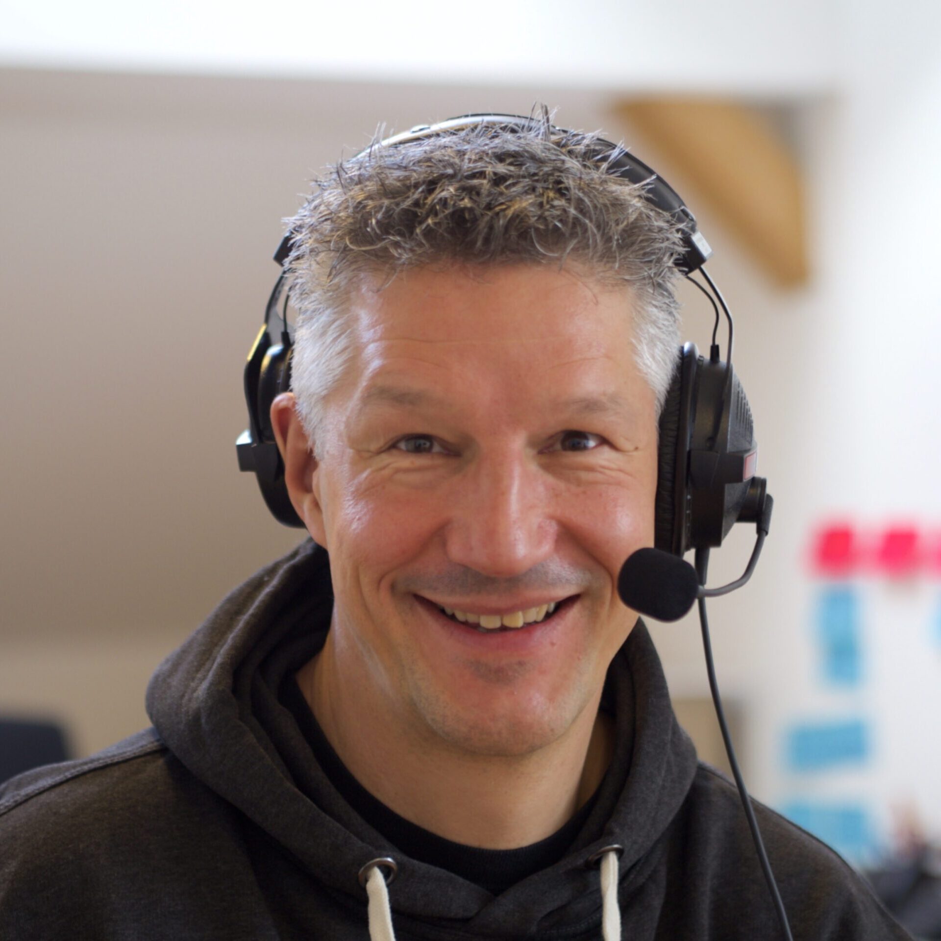 Photo of a German man wearing a headset with the microphone near his mouth. He has short, spikey grey hair and is wearing a dark coloured hoodie with white strings.