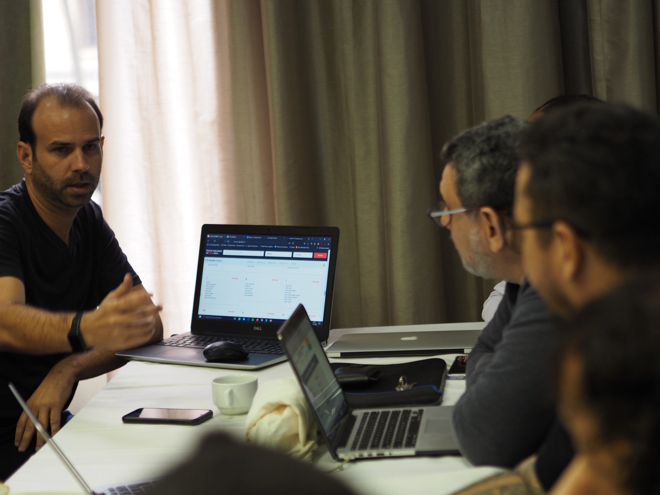 Photo of a man with a laptop open and three other people sitting on a row of tables looking at him and listening to what he's saying.