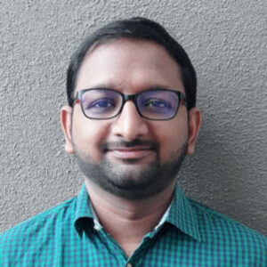 Photo of an Indian man wearing glasses smiling at the camera. He has a slight beard and is wearing a green shirt.