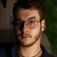 Photo of a Brazilian man looking at the camera. He has glasses on and half his face is in shadow. He has a slight beard, is wearing a thin necklace and a dark coloured shirt.