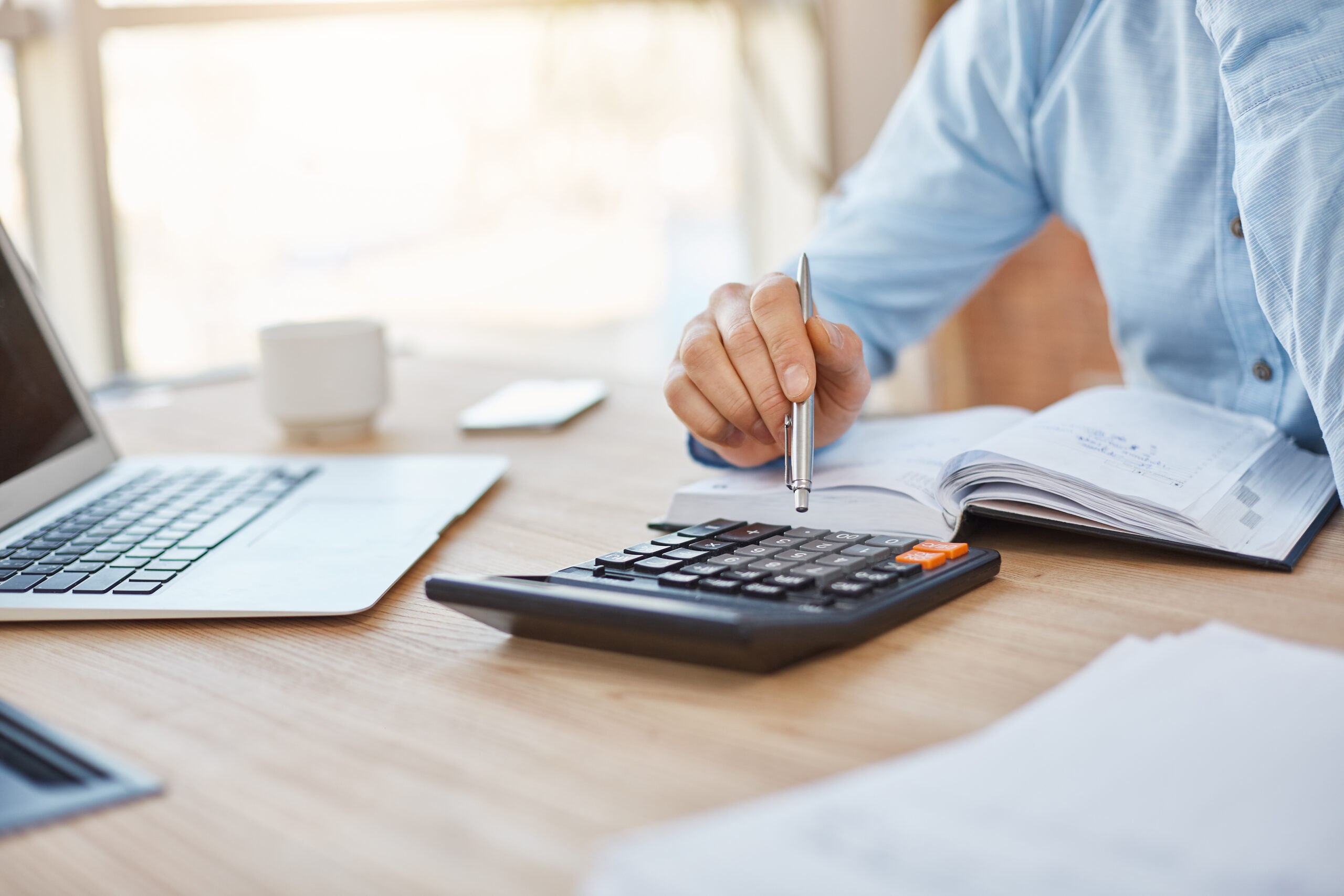 Close up detail of a person with a light blue shirt on, sitting in light office, checking company finance profits on calculator, writing down results in notebook.