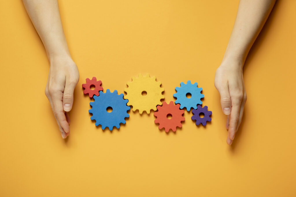 A yellow background with two hands a little way apart, palms facing each other, with six different size and colour cogs all connected to each other between the hands.