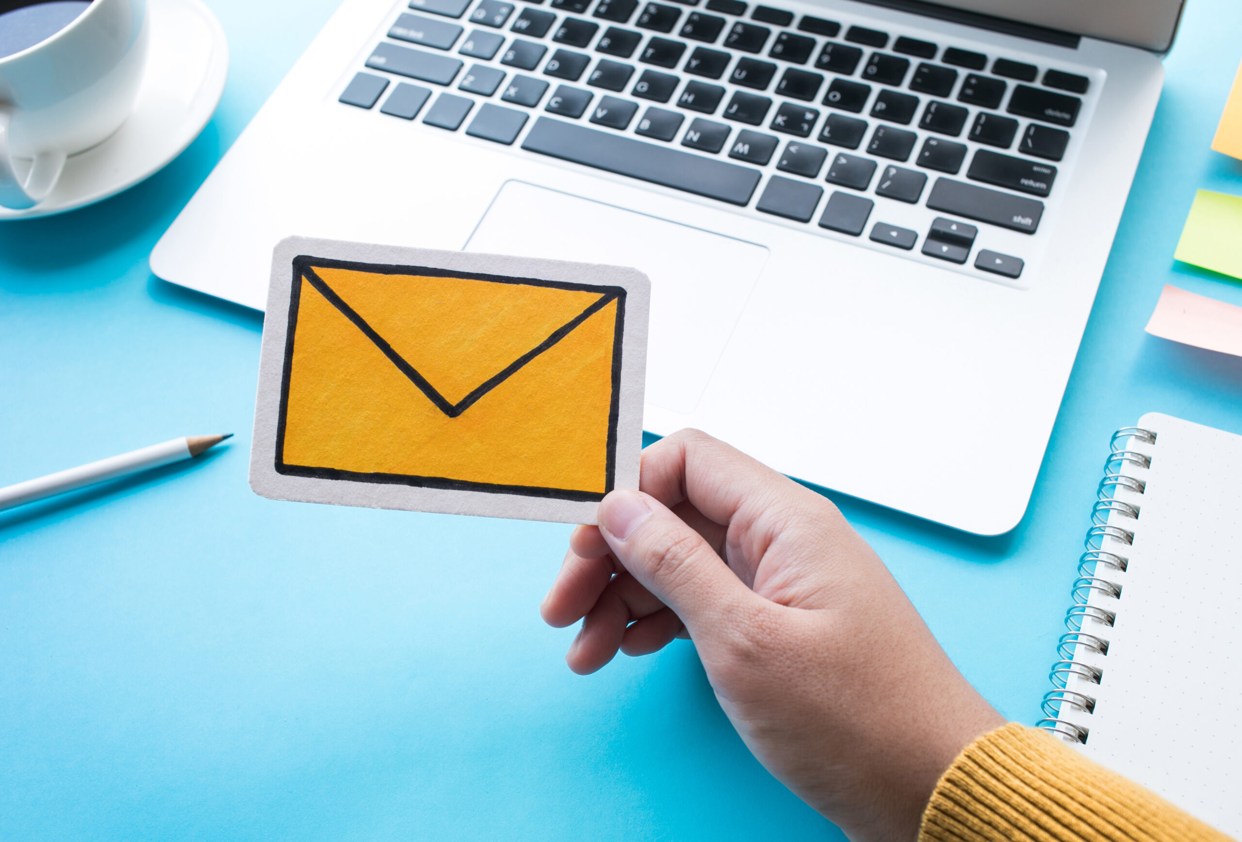 A laptop in the background with a hand in the foreground wearing a yellow woollen jumper holding a cut-out hand-drawn image of an envelope representing email.