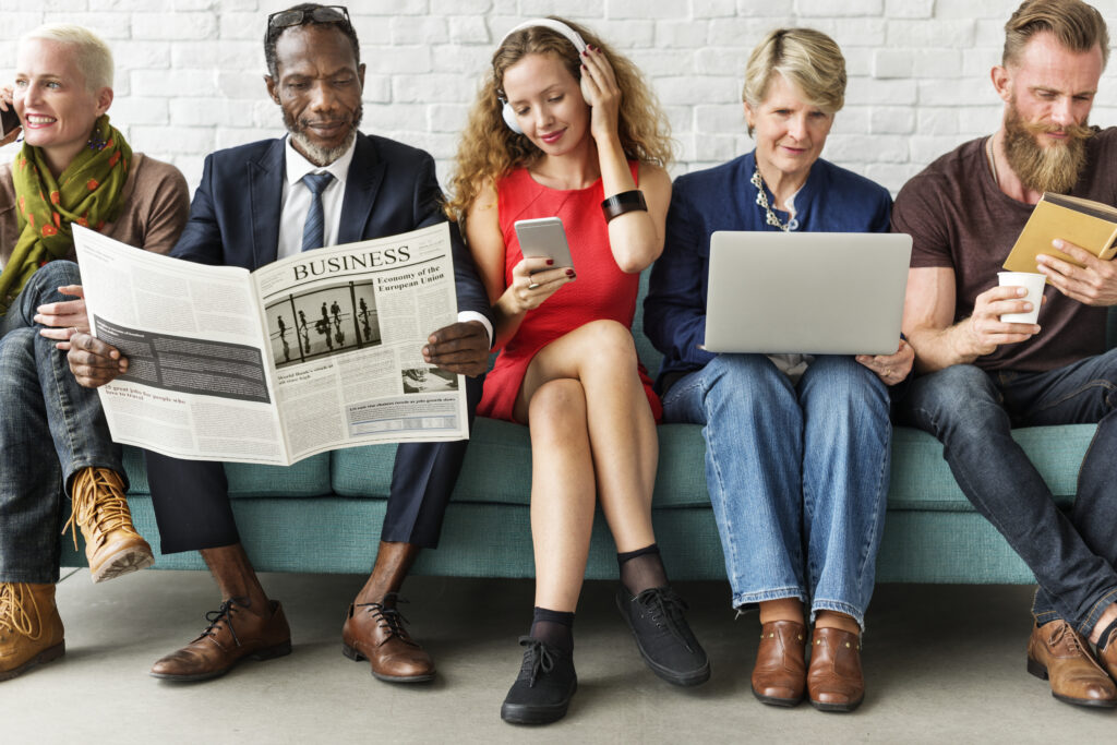 A group of diverse people is sitting together, one is on their phone, another reading a newspaper, another listening to music on a phone, another with a laptop on their knees and someone else reading a book.