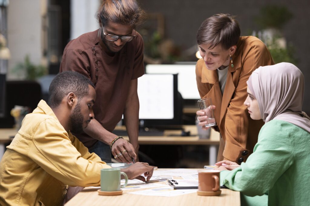 Photo of four people sitting around a table looking at charts. Two are standing and two are sitting. A woman on the left is wearing a pale pink hijab headscarf and a green long sleeved top, the woman standing has short dark hair and wears an orange coloured jacket. A man standing to the left has glasses and long, dark brown hair with lighter streaks and wears a brown side-buttoned kurta. The man sitting on the left wears a yellow long sleeved shirt with short black hair and a short beard.