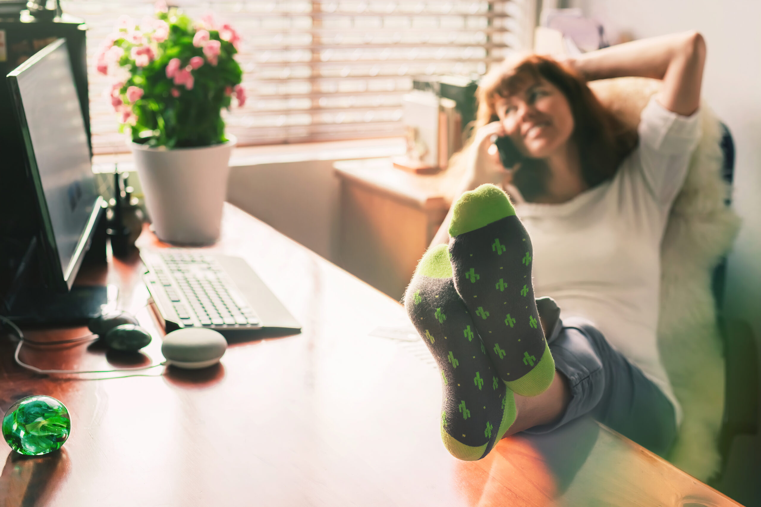 Blurred happy middle aged woman siting in a green chair in a home office, with her legs on the desk and speaking on a mobile phone.