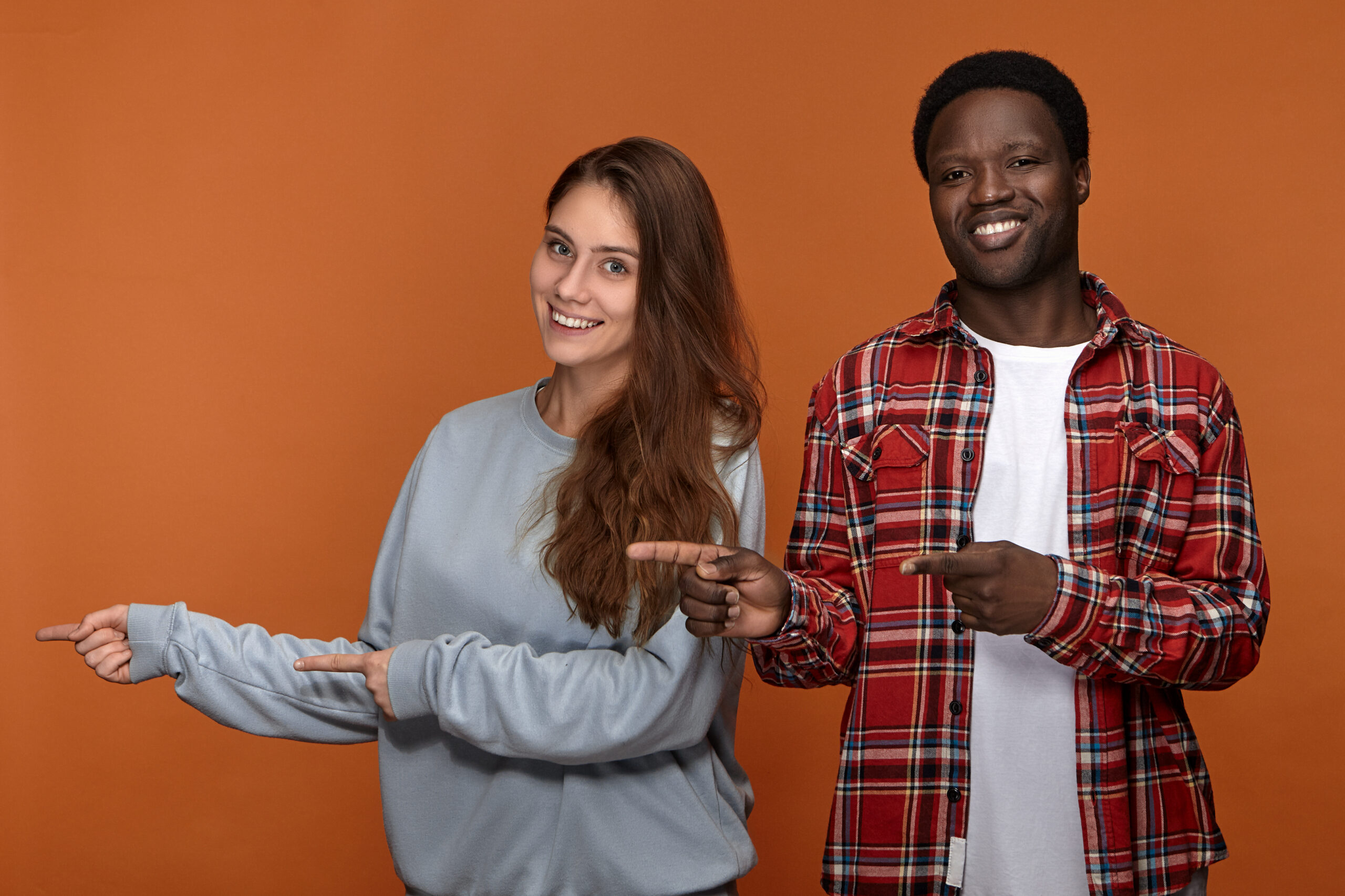 Picture of emotional overjoyed young Afro American guy in plaid shirt posing in studio with his adorable friendly looking Caucasian girlfriend, pointing fore fingers sideways and smiling broadly