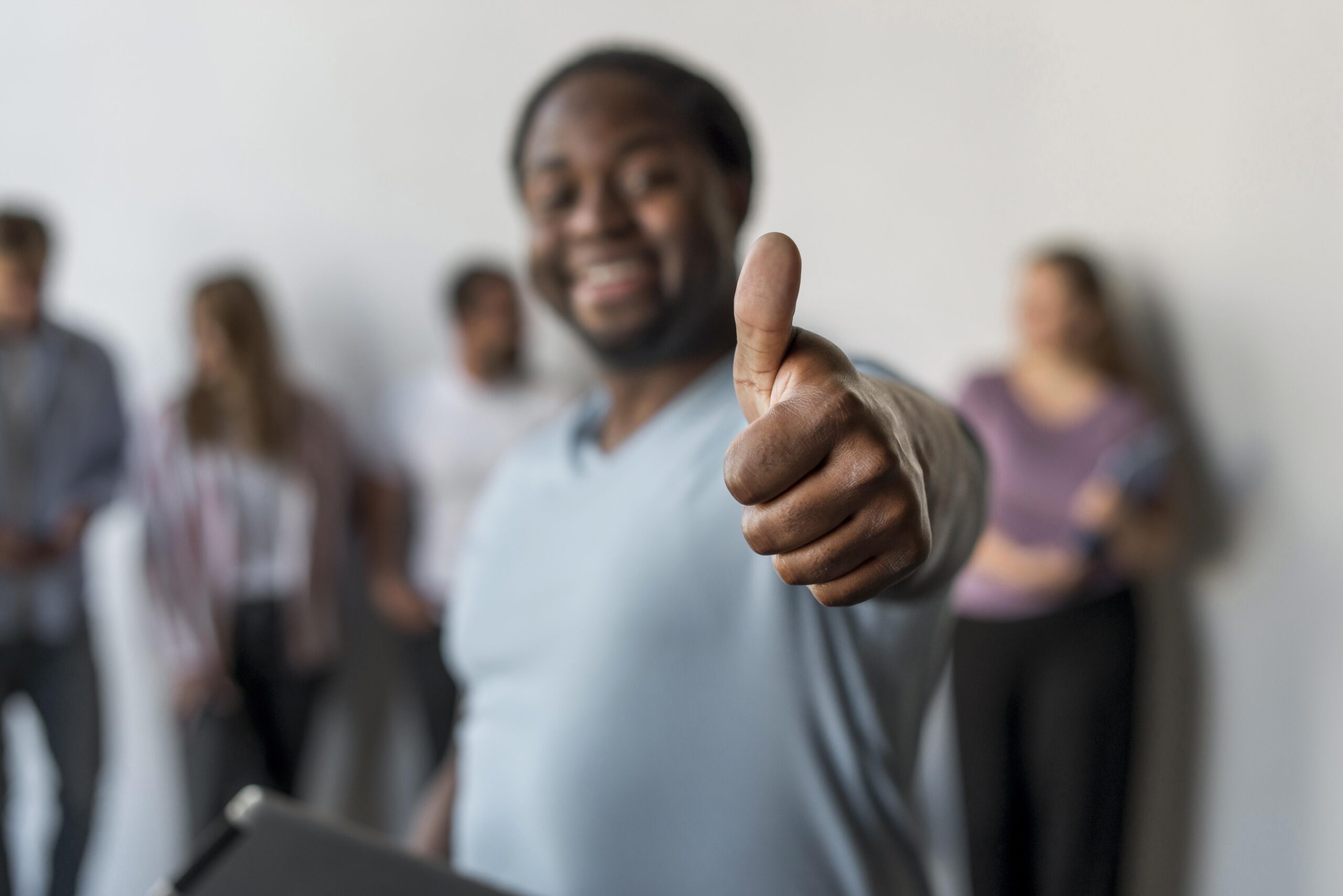 Photo of a black man wearing a pale blue tshirt facing the camera with his thumb up. In the background are a group of people but they are blurred and the main focus is the raised thumb in the foreground.