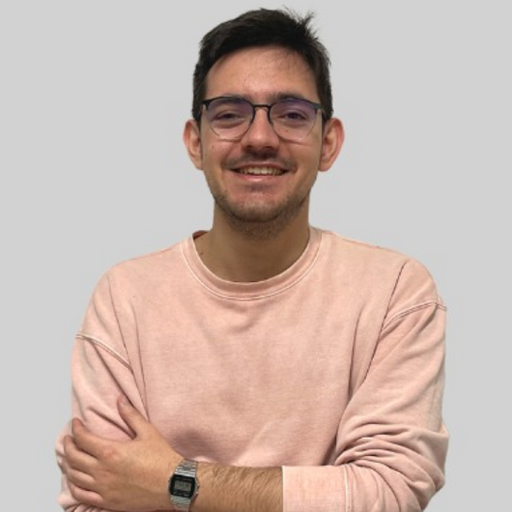 Photo of a portugese man with short dark hair wearing glasses and a light coloured jumper, standing to the camera with his hands folded in front of him.