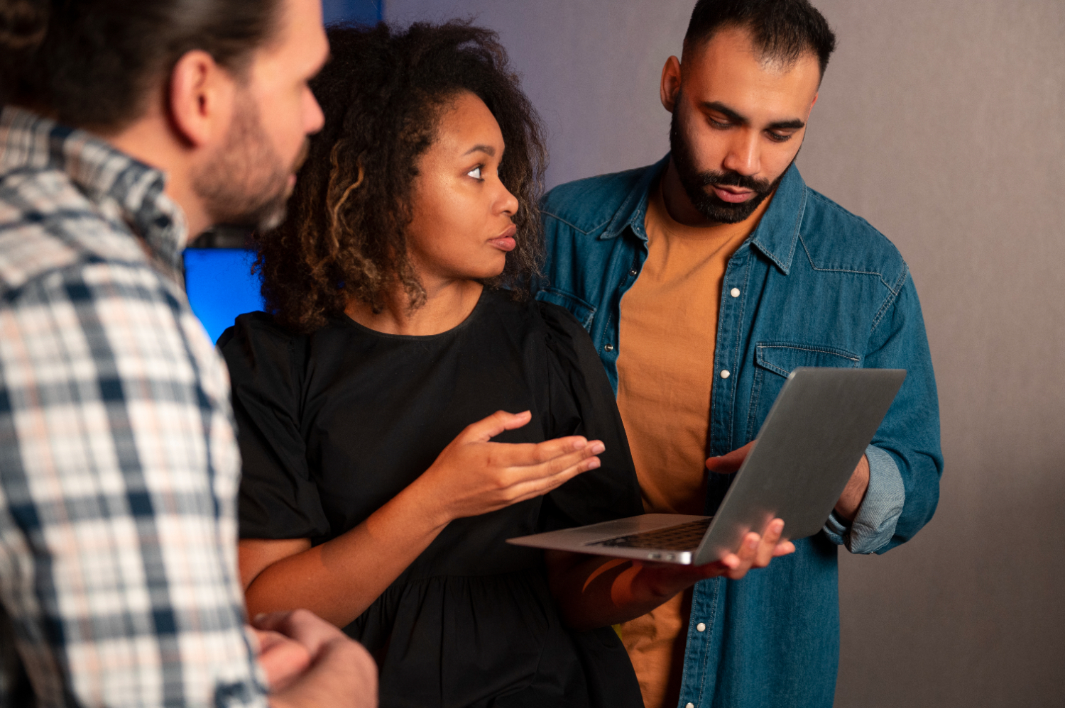 Three diverse people, two men and a woman, gathered around a laptop held by the woman in the middle, who points to the laptop screen.