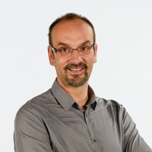 Photo of a white man with short hair and a beard and mustache wearing glasses. He has a dark, collared shirt on.