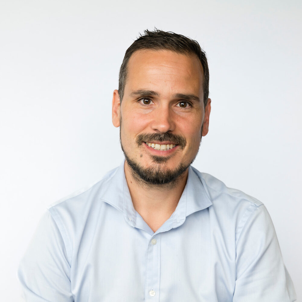 Photo of Dominique de Cooman, he's a white man with short black hair, a beard and a moustache. He's smiling to the camera and wearing a white open collared shirt.