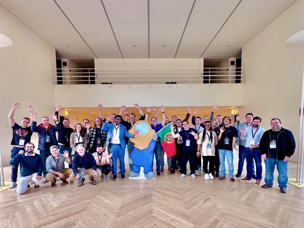 Photo of lots of Mauticians standing around a cardboard cutout of the Mautinaut holding a Portugal flag.