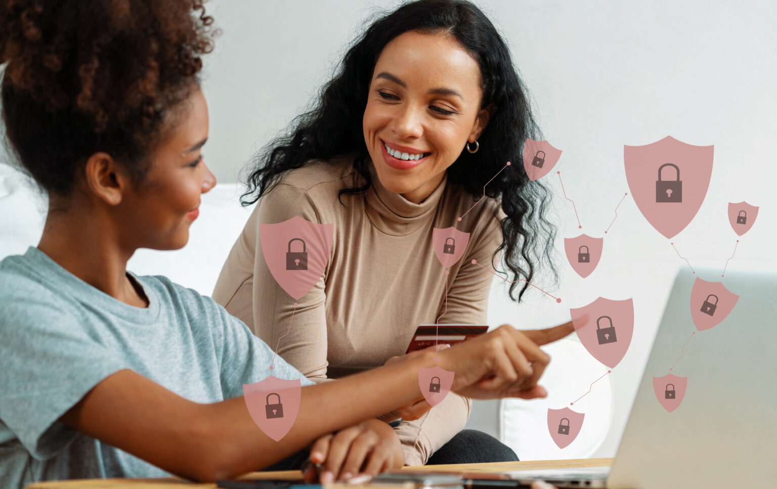 Black woman wearing a pale blue tshirt pointing at a laptop consulting another woman behind her with a hologram graphic interface of showing pale pink shields with a locked padlock in a darker colour.