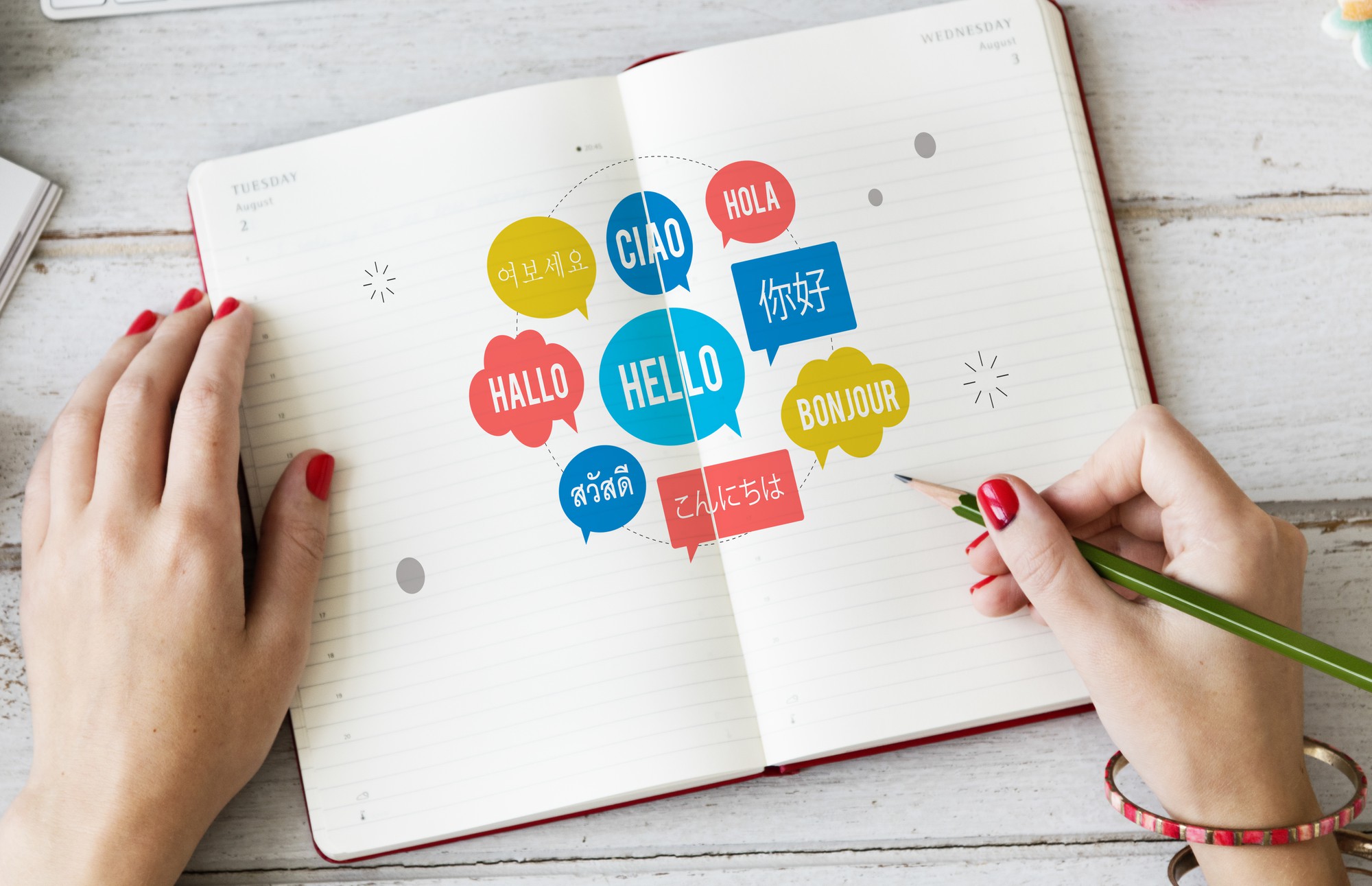 A photo of a notebook open on a table with the word hello written in speech bubbles in lots of different languages. Someone is holding the book open and has a pencil poised as if to write in the book.