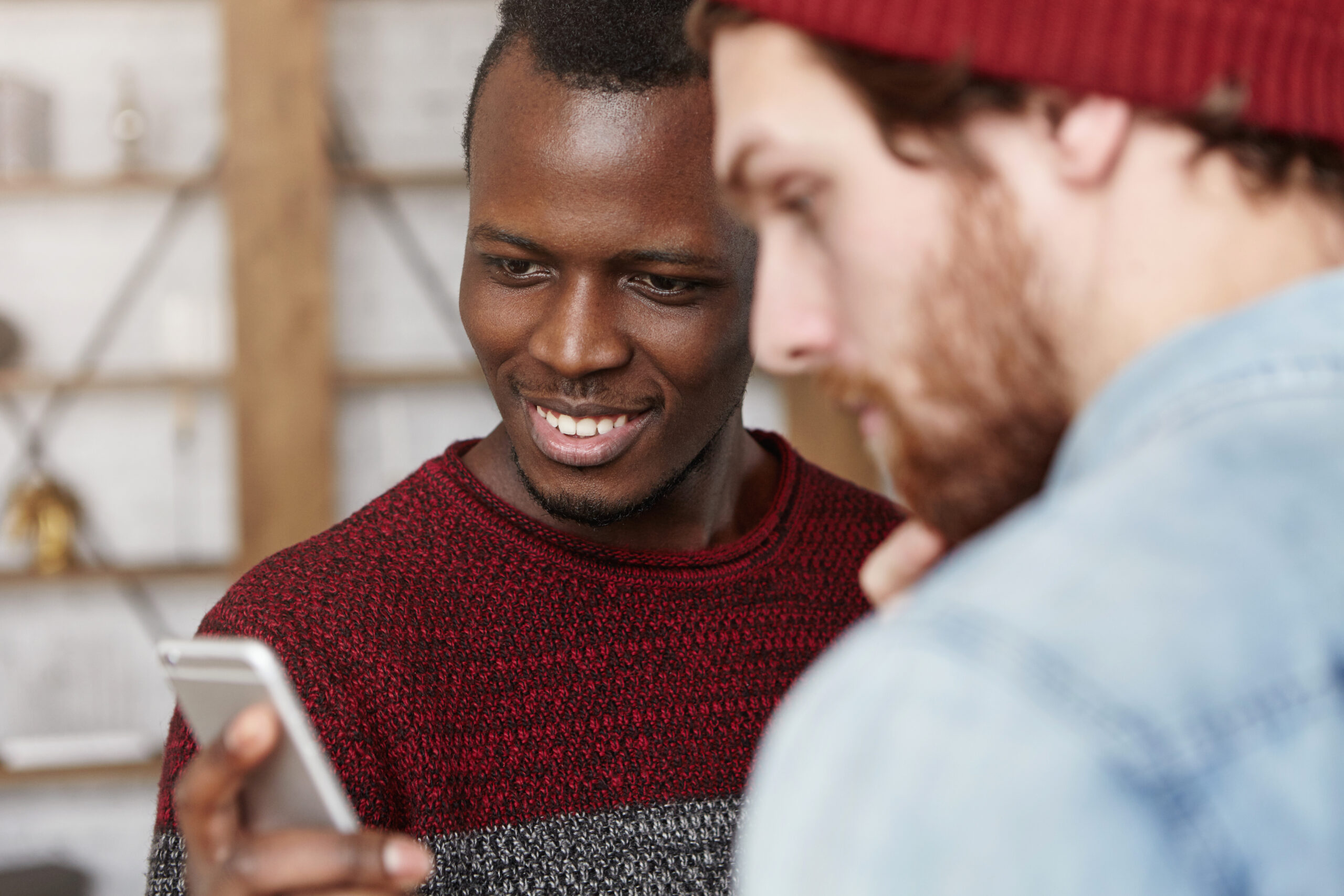 Excited happy young black man in sweater showing his fashionable white friend his mobile phone together at cafe.