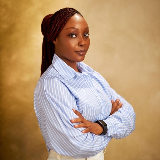 Photo of a black woman with long braided hair tied back, standing against a mottled brown background with her arms folded, wearing a light blue coloured shirt and white trousers, with a black watch on her wrist.