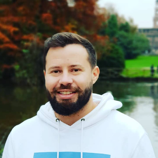 Photo of a white man with short dark hair, a full beard and mustache, wearing a white hoodie.