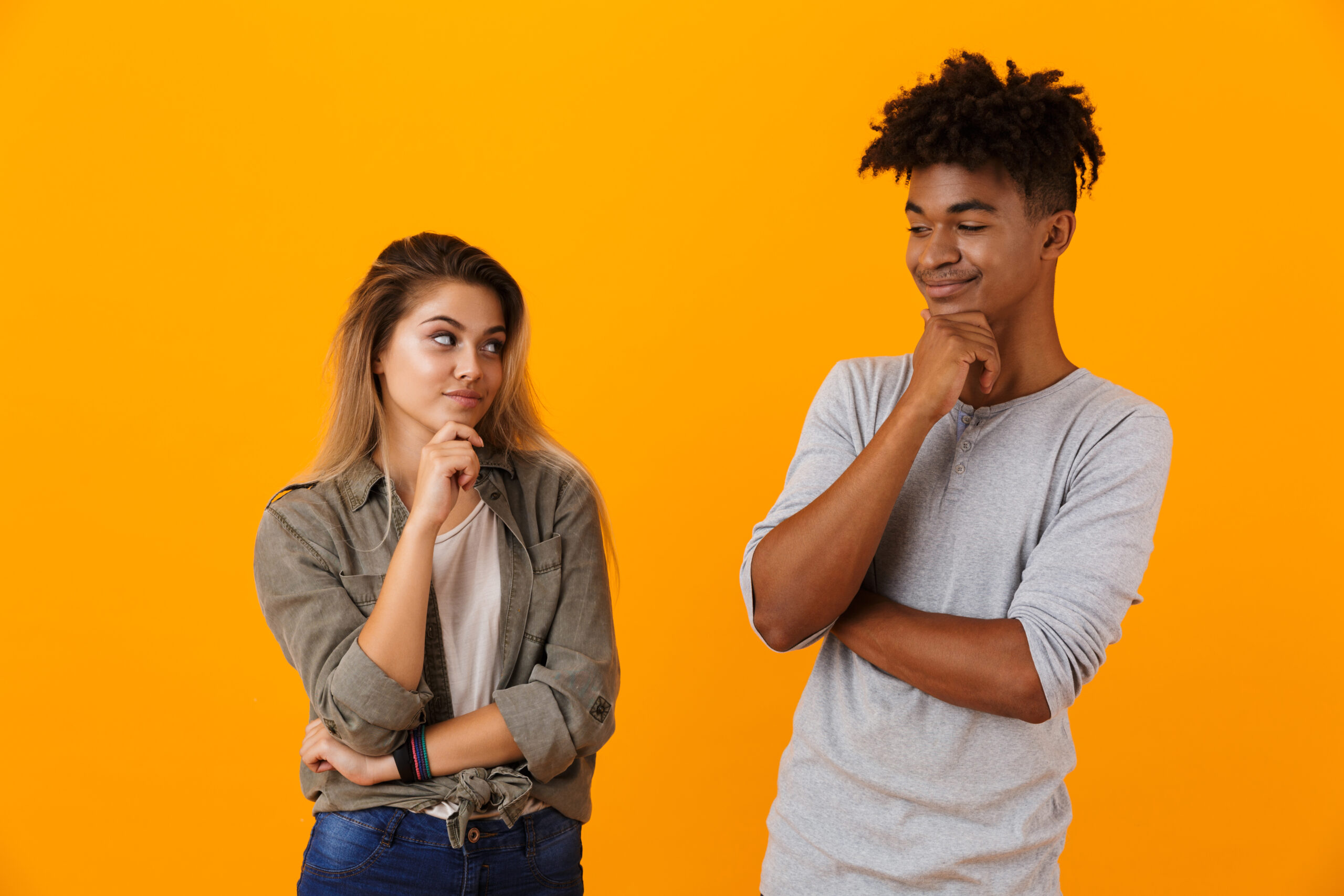 Portrait of a smiling pensive multiethnic couple standing isolated over yellow background, looking at each other