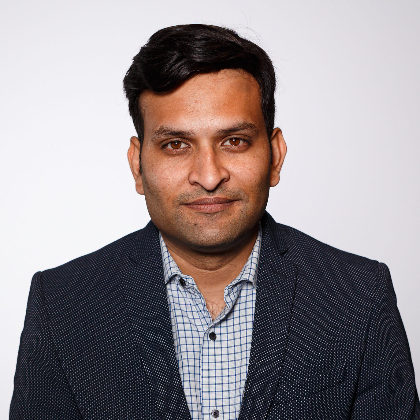 A photo of Prateek, he's an Indian man, clean shaven with dark mid-length hair wearing a black blazer and a checked white and blue shirt. He is smiling at the camera and is photographed against a white background.