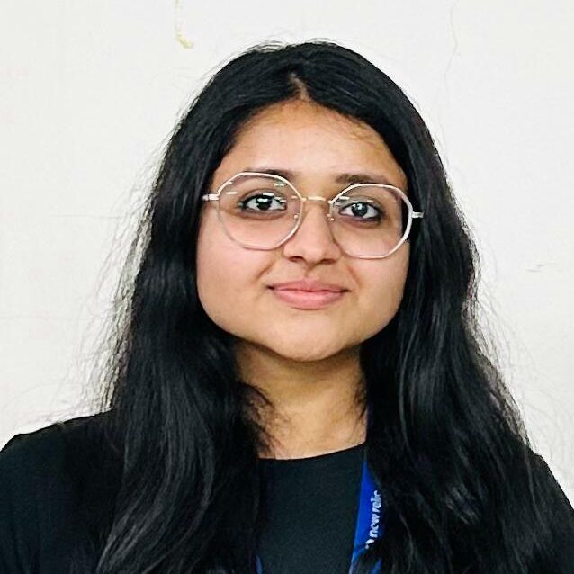 Photo of an indian woman with long dark hair worn loose, she is wearing glasses and dark clothes with a blue lanyard.