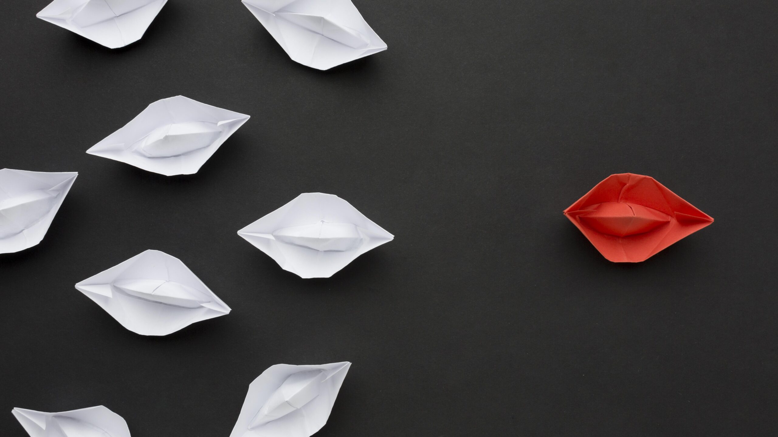 Image of a red origami boat in front of multiple white origami boats on a black background.