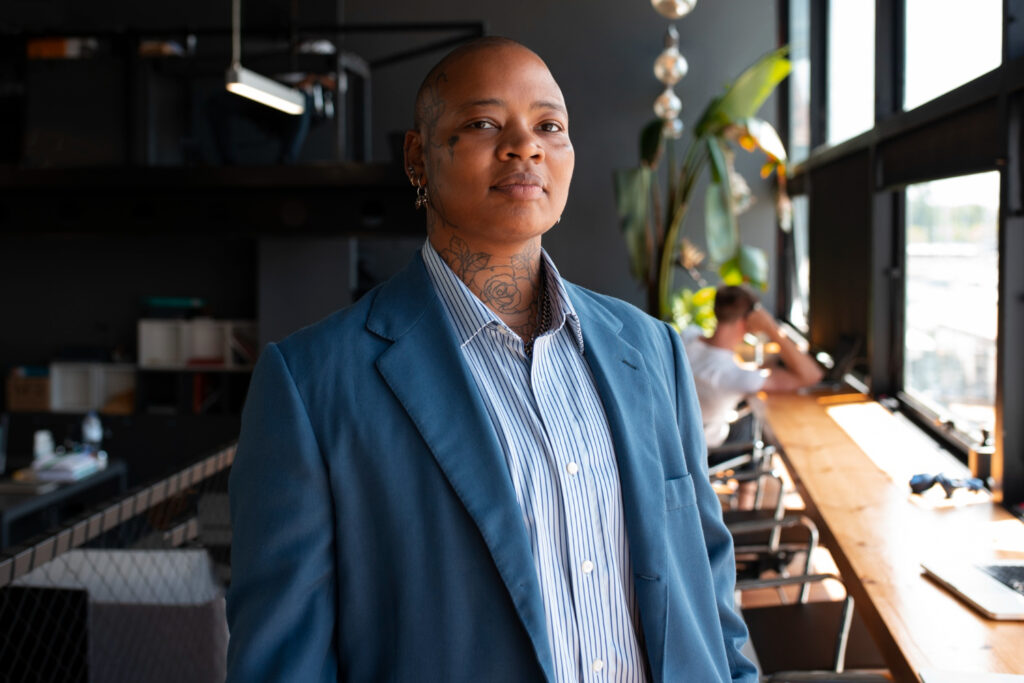 A photo of a black person standing beside a wooden bench in what looks like a workplace, they have a shaved head and wear an unbuttoned blue and white striped collared shirt and a blue suit jacket. They have earrings in their ears and tattoos are visible on their neck and back of their head.