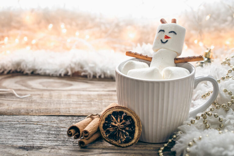 Photo of a mug containing hot chocolate with marshmallows, and some of the marshmallows have been made into the shape of a snowman with a face drawn on and hands and ears added. There are oranges and cinnamon on the table beside and behind is a blurred image of festive lights.