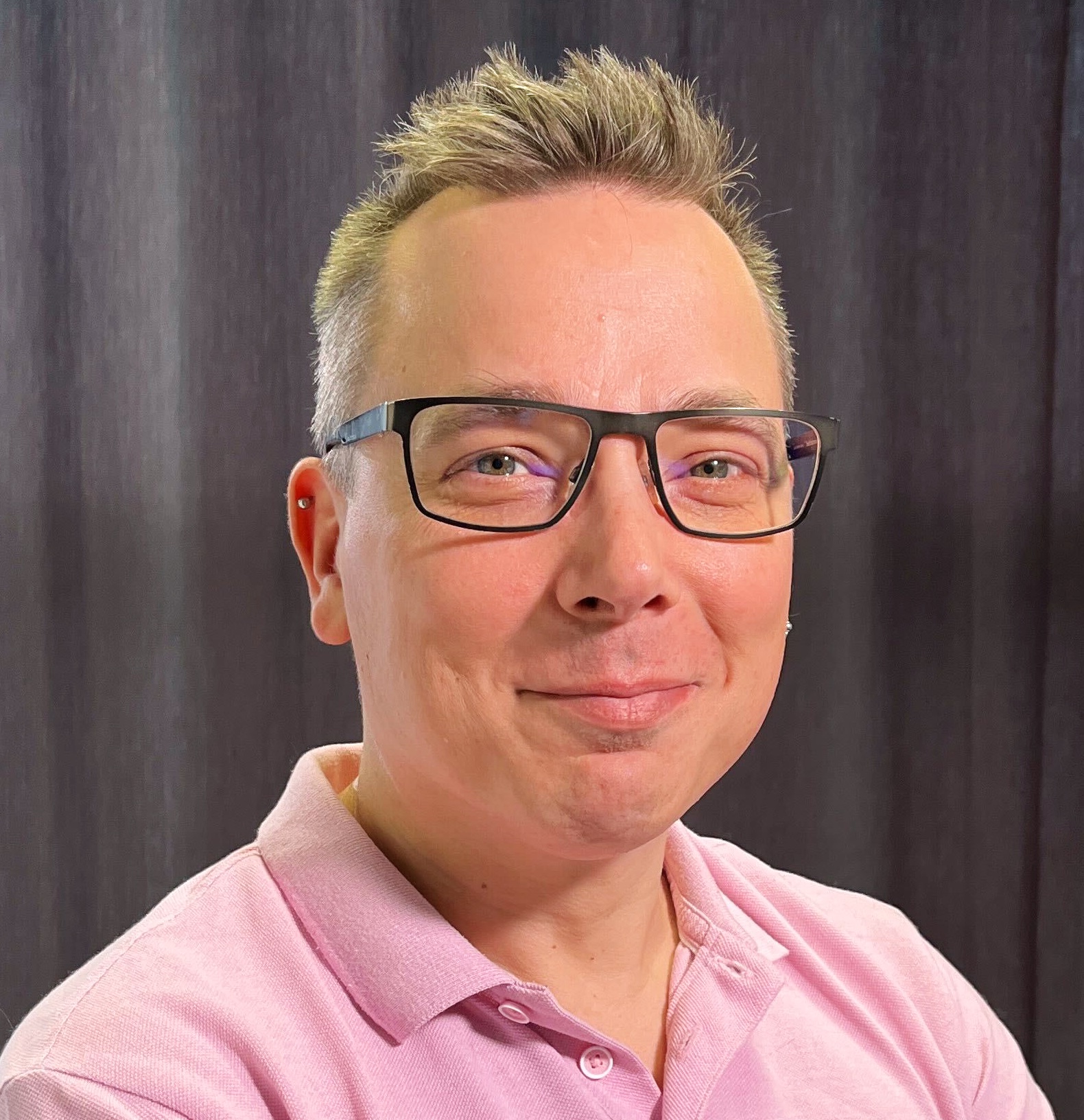 Photo of a white man wearing glasses with short fair hair spiked at the front. He's wearing a pink collared shirt with the top buttons undone.