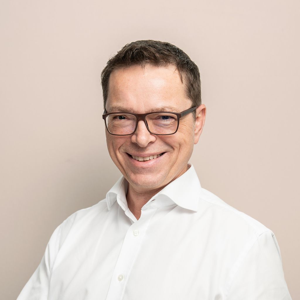 Photo of a white man with short dark hair wearing glasses. He's got a white unbuttoned collared shirt on.