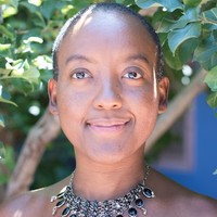 Photo of a black woman with a shaved head wearing a decorative necklace with dark stones. She's standing in front of a tree.