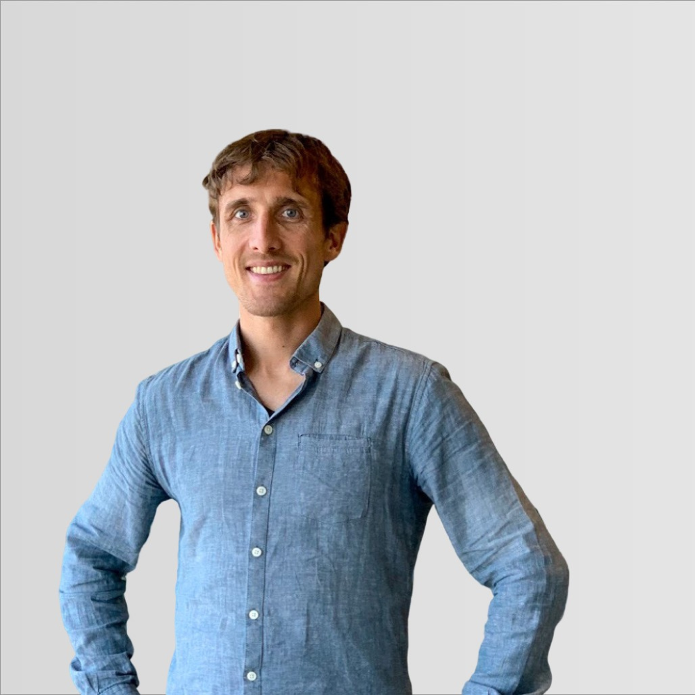 Photo of a white man with short light brown hair. He's got his hands on his hips and he's wearing a pale blue collared shirt with the top buttons undone