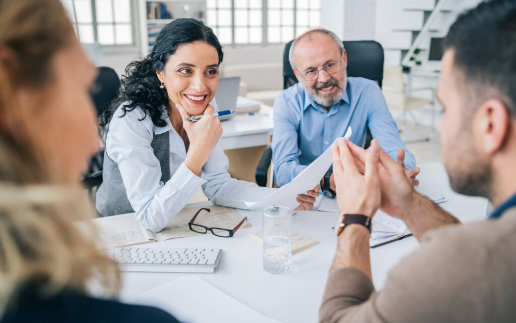 Photo in and office environment of a woman and a man sitting to a table discussing something. The woman is holding a piece of paper facing the camera and there is someone sitting behind the camera view with their elbows on the table, palms together, facing the other two.