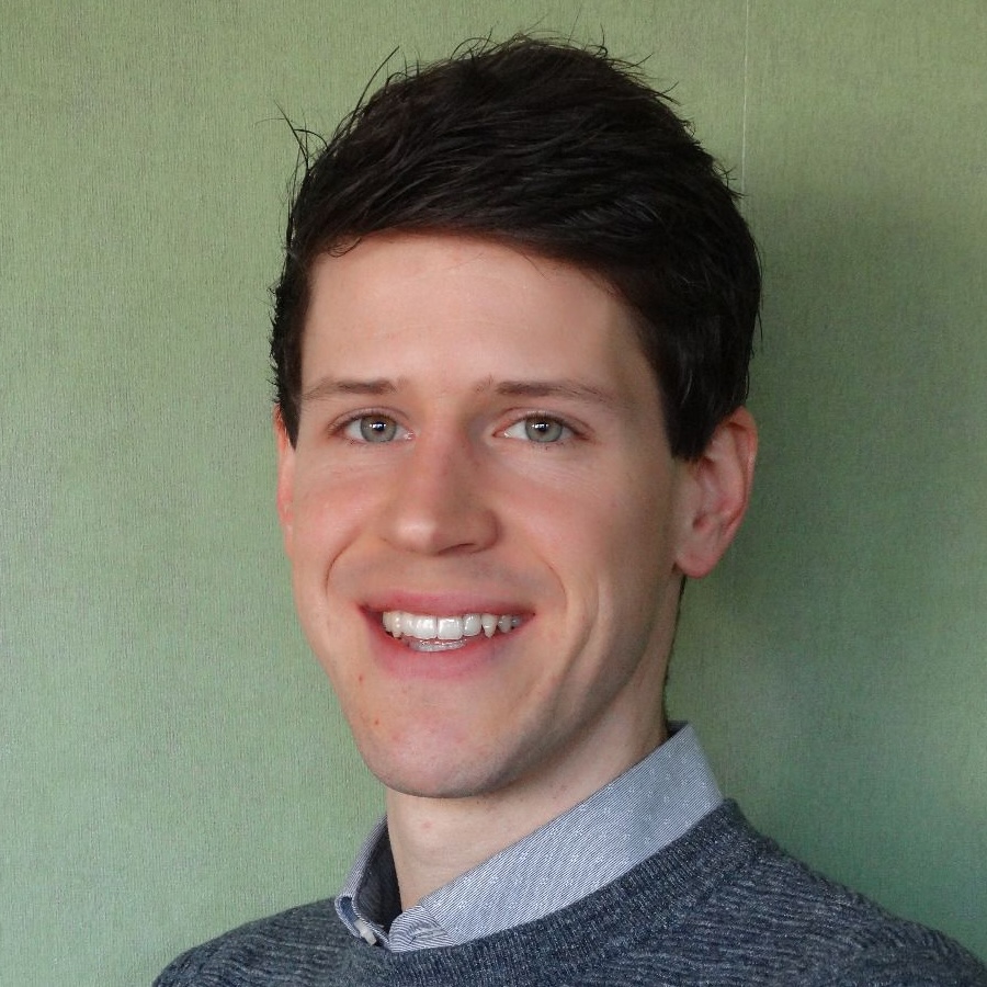 Photo of a white man with short dark hair wearing a light grey collared shirt and a dark grey jumper, pictured against a green background.