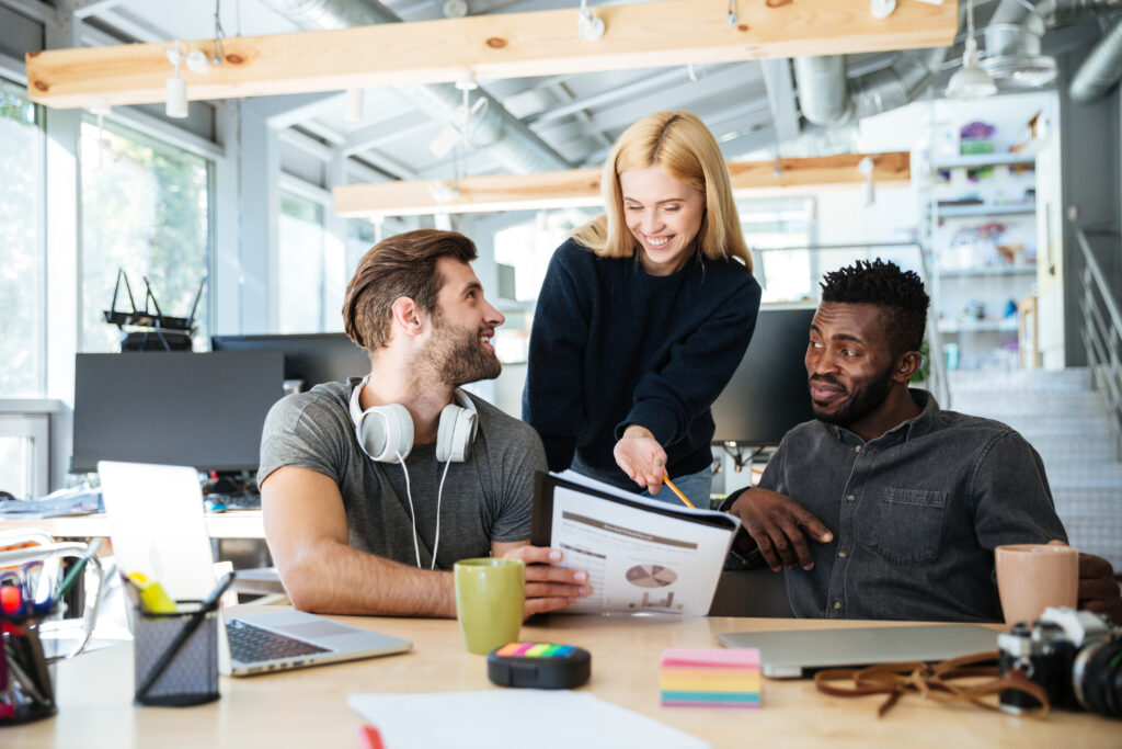 Image of happy young colleagues in office coworking using laptop computers and talking with each other. Looking aside.