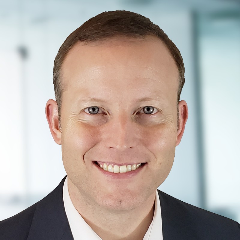 Photo of a white man with short cut dark hair, smiling at the camera wearing a white shirt and a dark suit jacket.