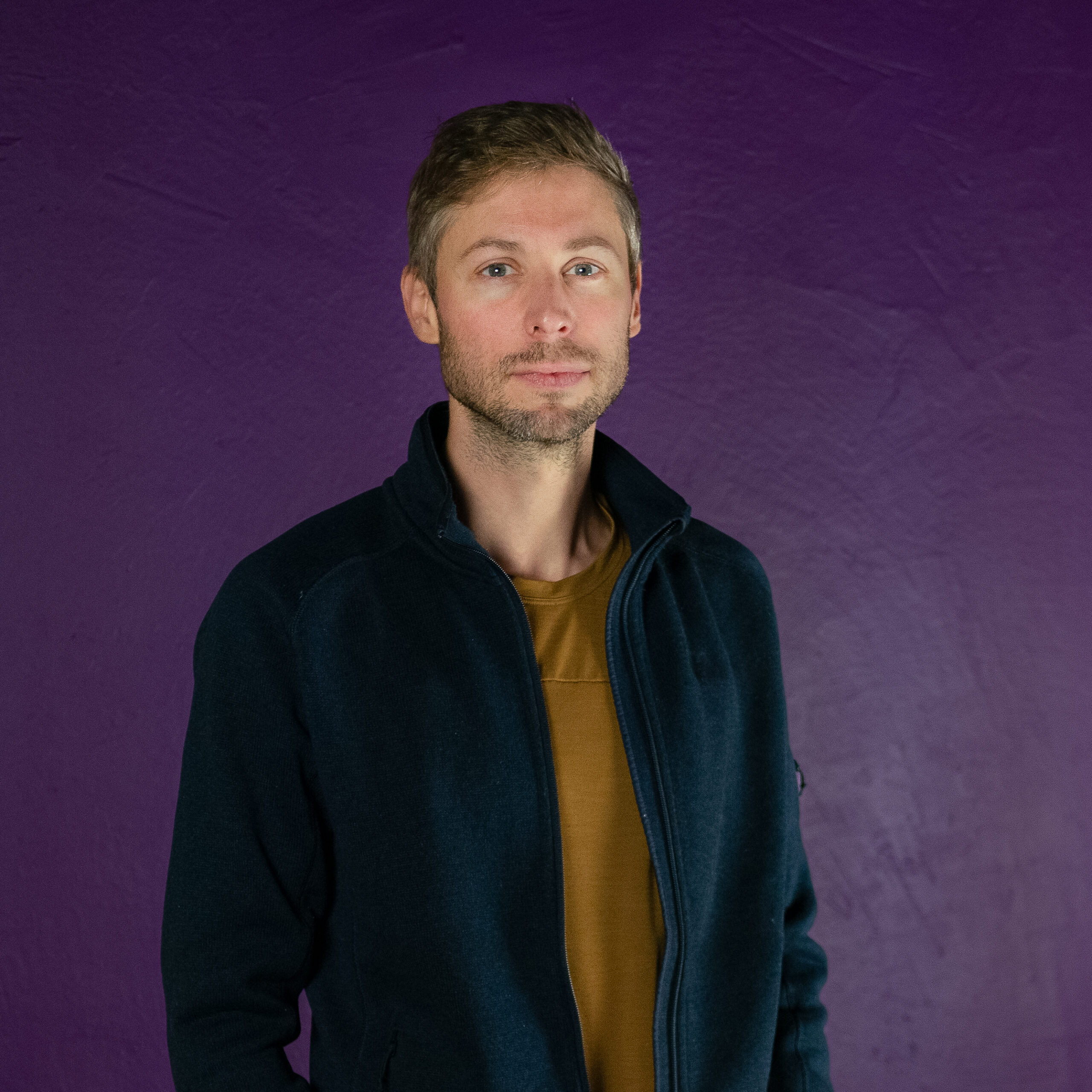 Photo of a white man with short, spiked light brown hair and a beard and moustache standing against a dark purple background. He's wearing a dark black jacket and a light brown coloured shirt.