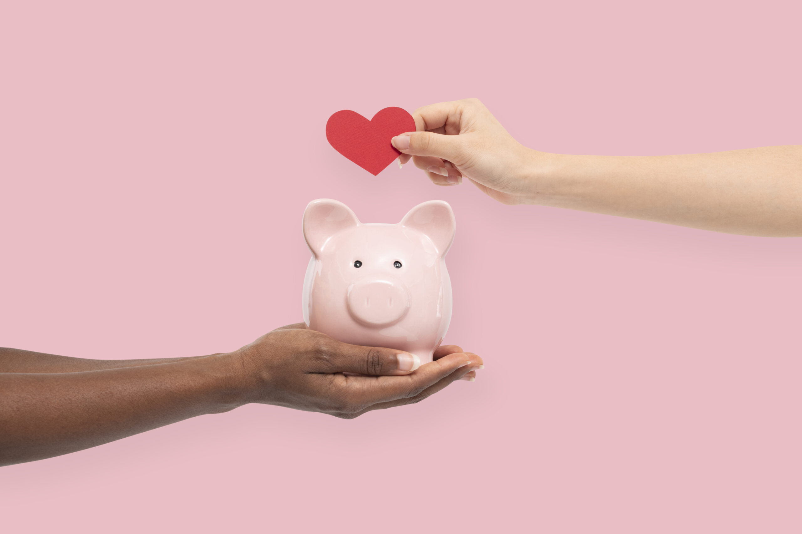 Photo of a pink background with a hand holding a piggie bank and another dropping a red heart into it.