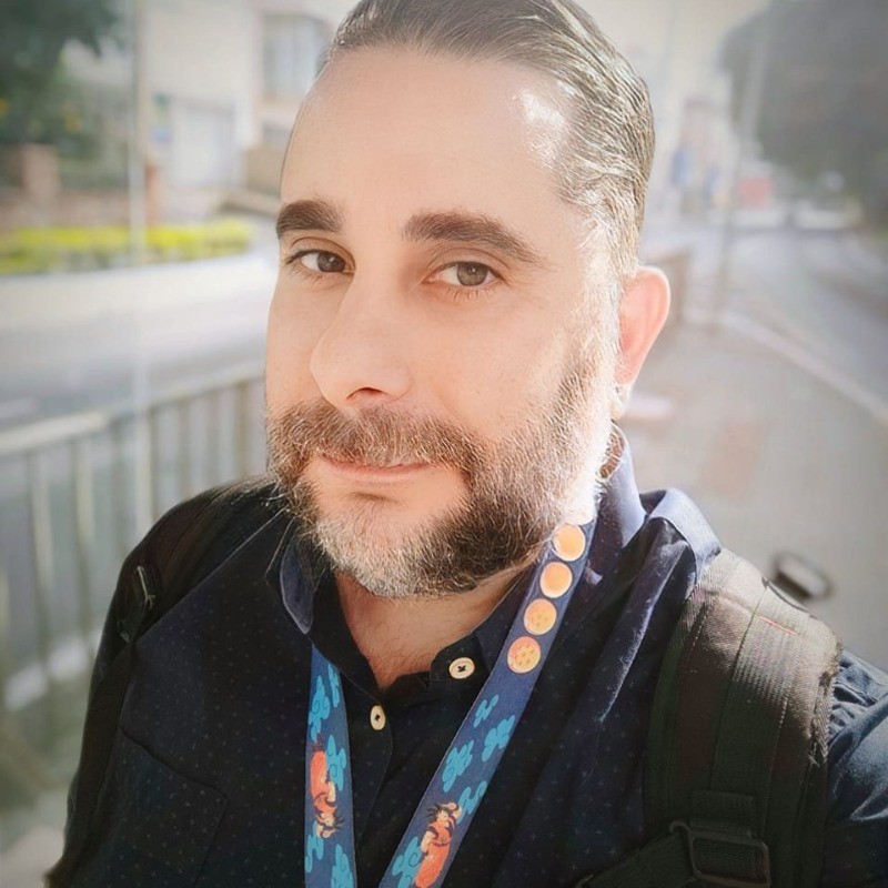 Photo of a Brazilian man with long hair tied back, and a moustache and beard. He's wearing a black shirt and a colourful lanyard, and seems to be pictured beside a main road.