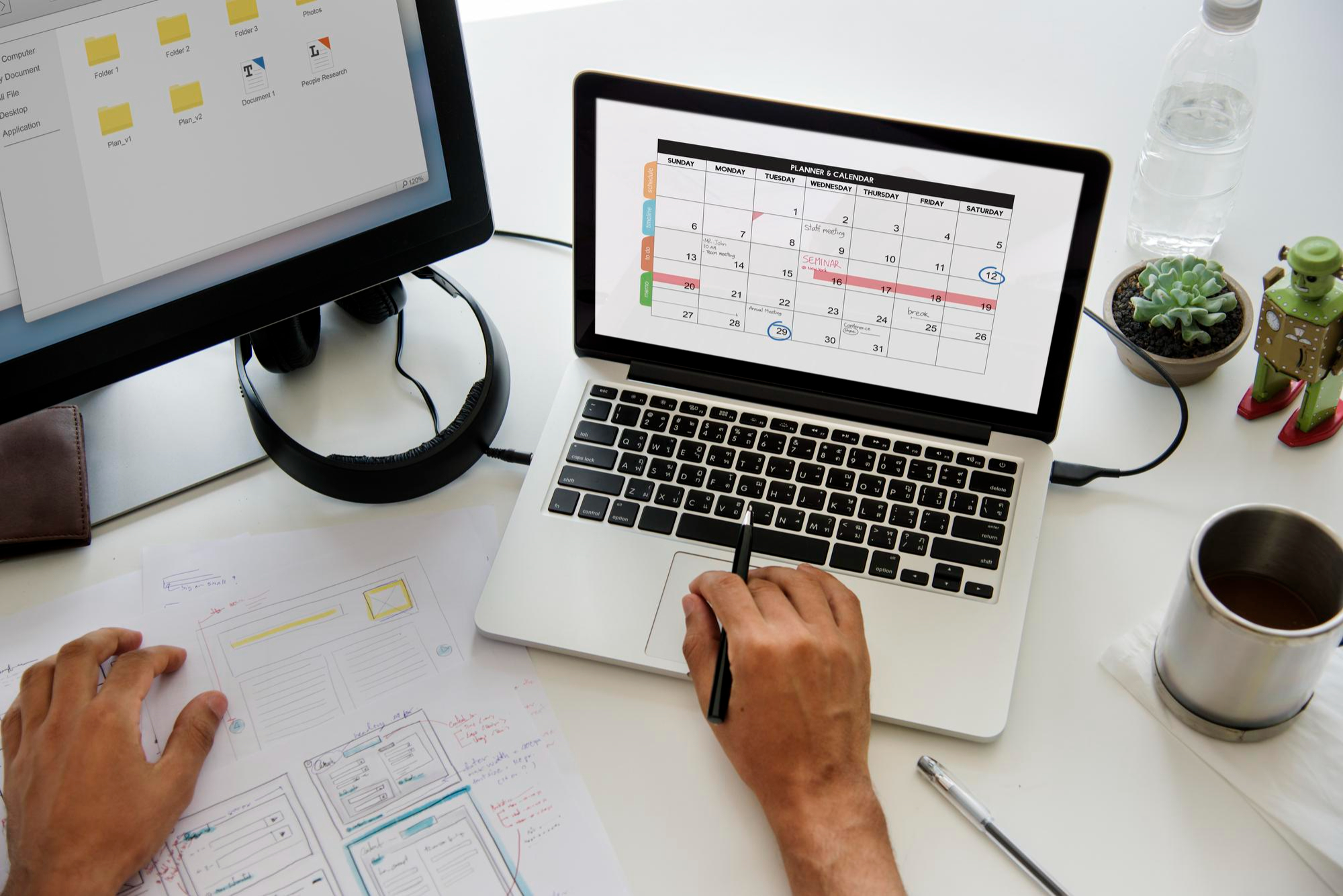 Image of a person using a calendar on a laptop screen with a monitor nearby and sketches on a table beside them.
