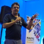 Photo of a Brazilian man standing on a stage wearing a dark tshirt with Mautic marketing software on the front. He's holding a microphon and the background is illuminated in blue, with a speaker behind him and a popup with Mautic Conference to the right. He's wearing jeans and has glasses with short dark curly hair.
