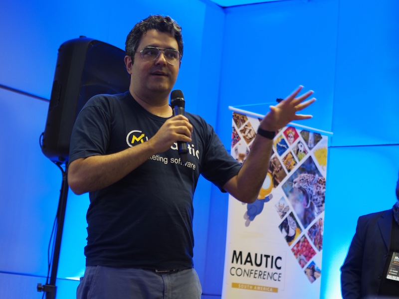 Become a Mautic Ambassador 6 Photo of a Brazilian man standing on a stage wearing a dark tshirt with Mautic marketing software on the front. He's holding a microphon and the background is illuminated in blue, with a speaker behind him and a popup with Mautic Conference to the right. He's wearing jeans and has glasses with short dark curly hair.