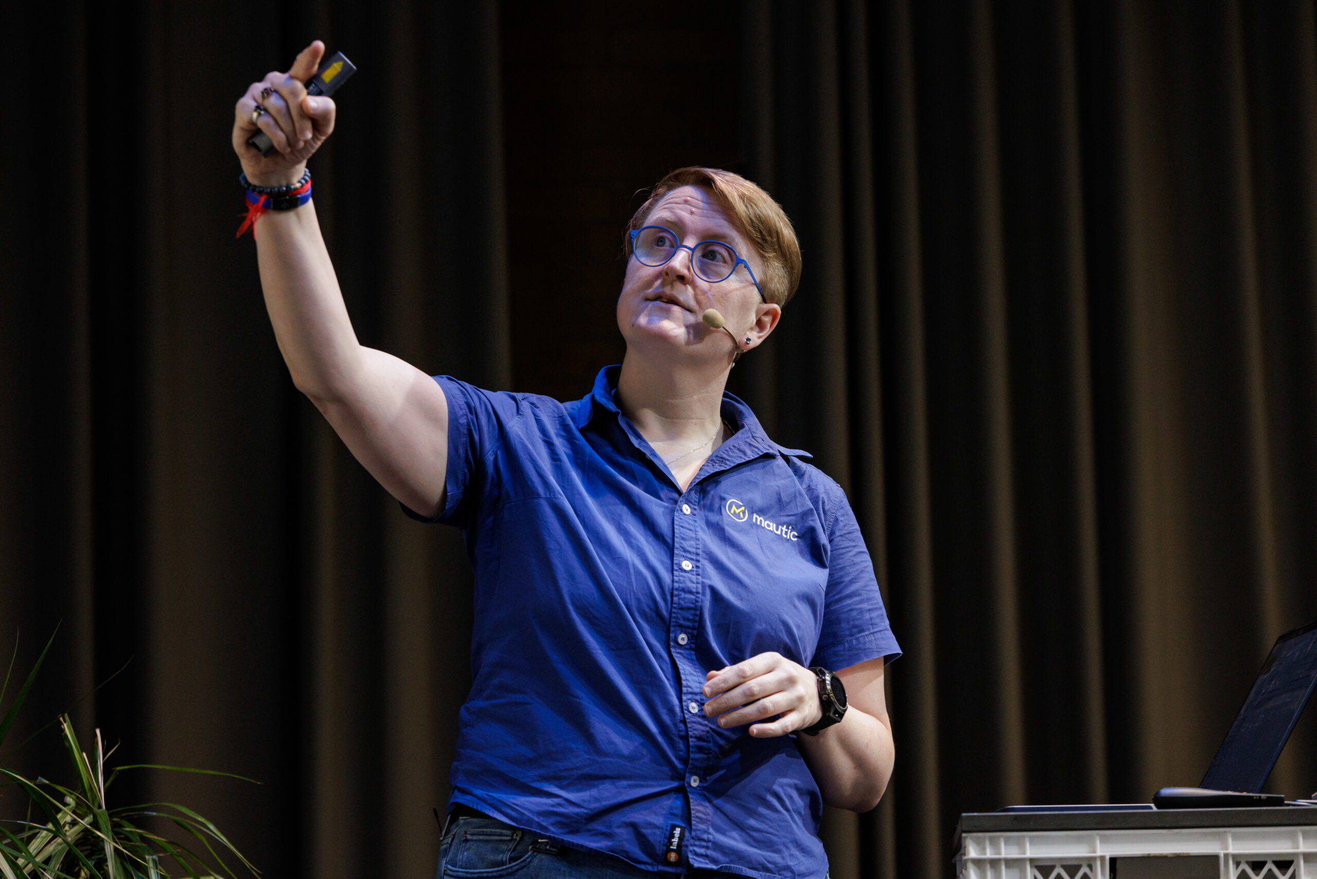 Become a Mautic Ambassador 5 Photo of a white woman with short blonde hair on the stage at a conference wearing a blue collared shirt with short sleeves. She's holding a remote presenter in her left hand and pointing to a screen with the right hand.