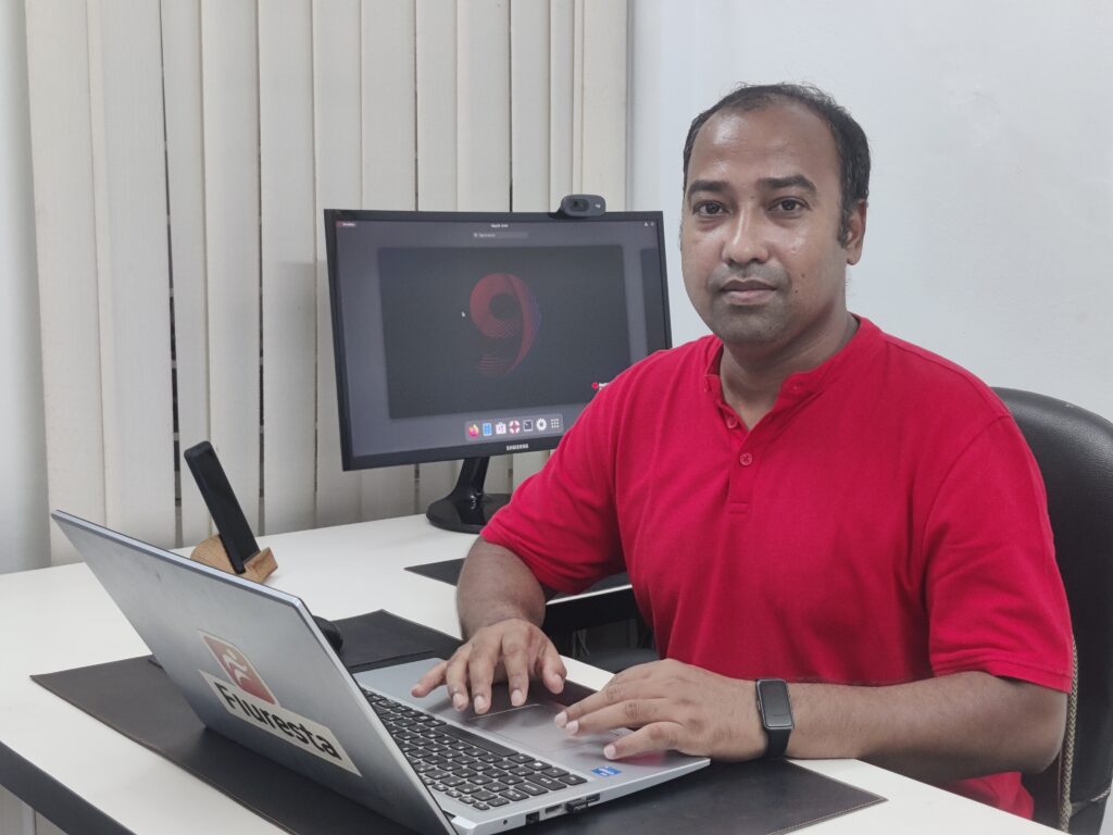 Photo of Ashique Rahman, a Bangladeshi man sitting at a desk with a laptop in front of him, wearing a red v-neck shirt.