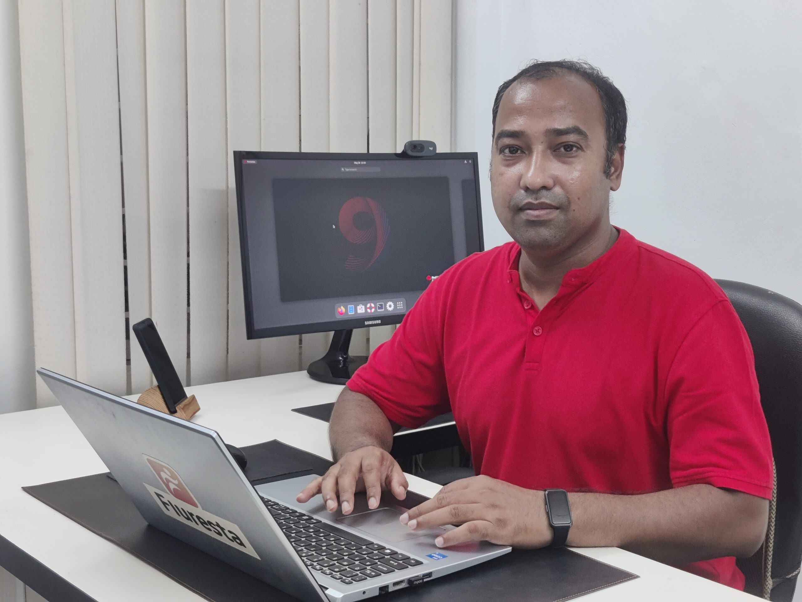 Photo of Ashique Rahman, a Bangladeshi man sitting at a desk with a laptop in front of him, wearing a red v-neck shirt.